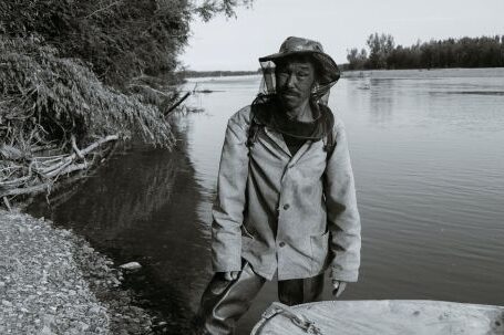 Fishing - Black and white of calm mature ethnic fisherman in anti mosquito head net hat standing in shallow water of lake near wooden boat and looking at camera