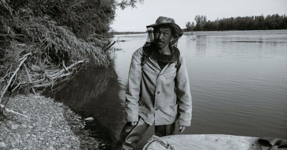 Fishing - Black and white of calm mature ethnic fisherman in anti mosquito head net hat standing in shallow water of lake near wooden boat and looking at camera